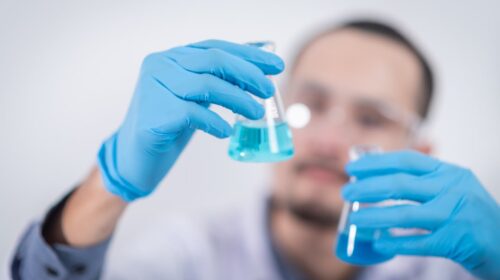 Ein Labormitarbeiter sieht sich mit einer blauen Flüssigkeit gefüllte Gefäße an.